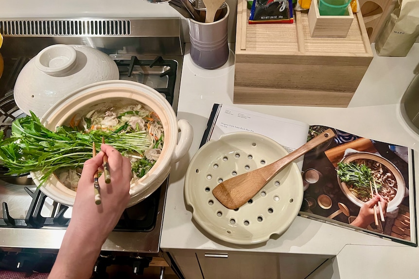 A hand placing greens in a claypot on a stove using chopsticks, mimicking an image from a cookbook.