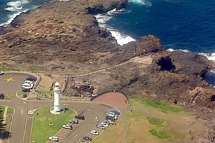 An aerial shot of a blowhole, headland, lighthouse and parking lot.