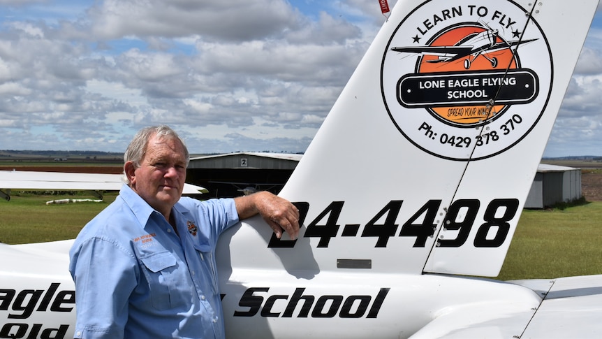 Trevor Bange stands next to a light plane. he wears a light blue shirt and his arm is resting on the side of the plane.