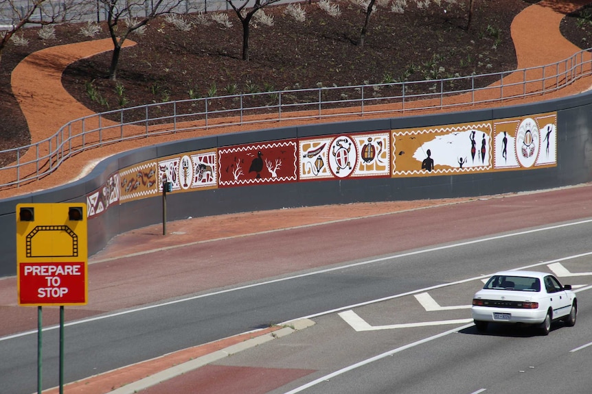Long panel of aboriginal art at the entrance to tunnel with car driving past, WA