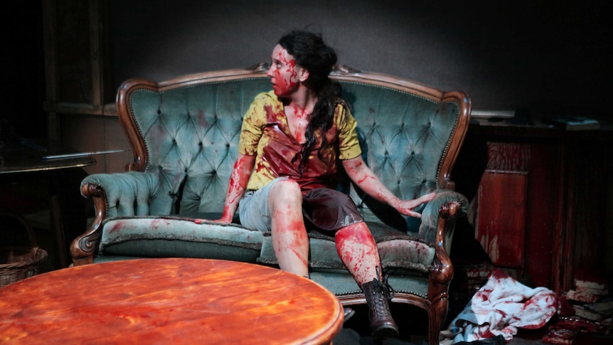 A woman covered in fake blood sits on a green couch and looks off camera.