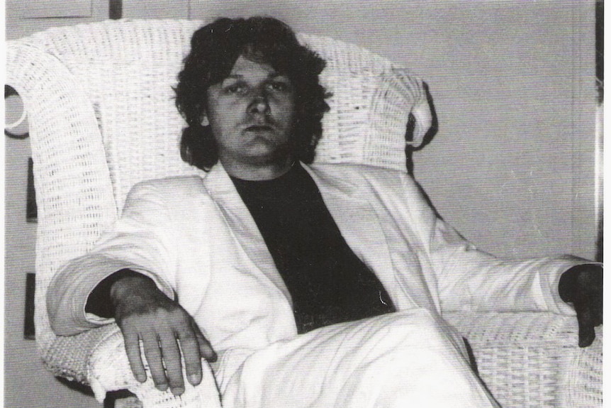 A young man with wavy hair, wearing a white suit, sits in a high-backed wicker chair.