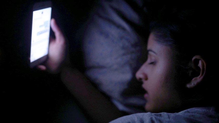 Somya holds a phone with her eyes closed in bed.