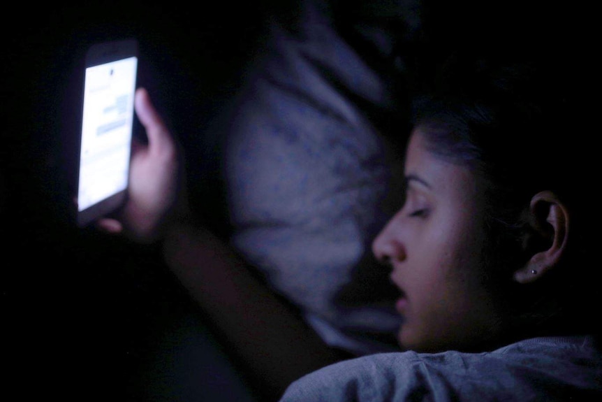 Somya holds a phone with her eyes closed in bed.