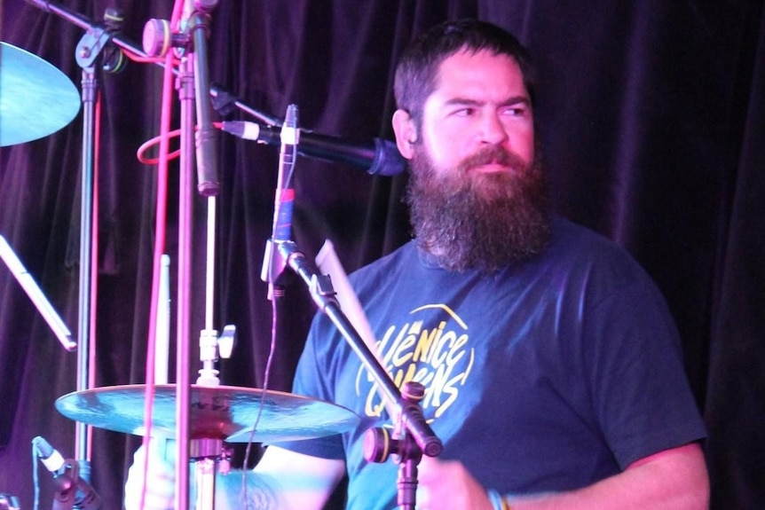 A man with a large beard plays the drums and looks off to the side.