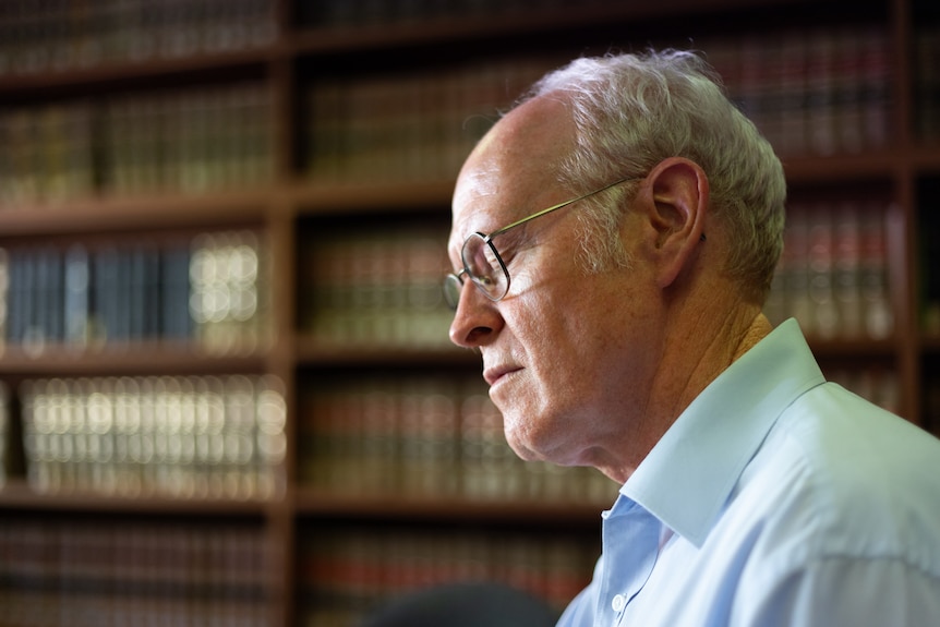 Side profile of a man wearing glasses and a blue shirt. A bookshelf is in the background.