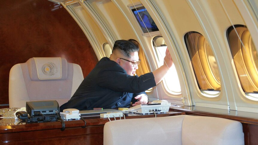 Kim Jong-un looks out an airplane window while sitting at a desk on a plane.