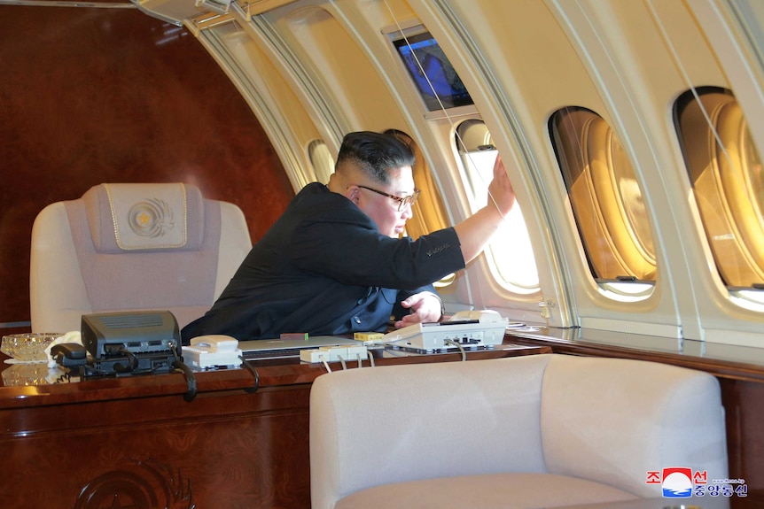 Kim Jong-un looks out an airplane window while sitting at a desk on a plane.