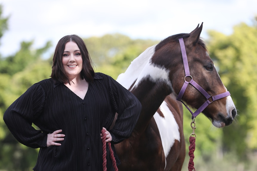 A woman with a black dress standing in front of her horse.