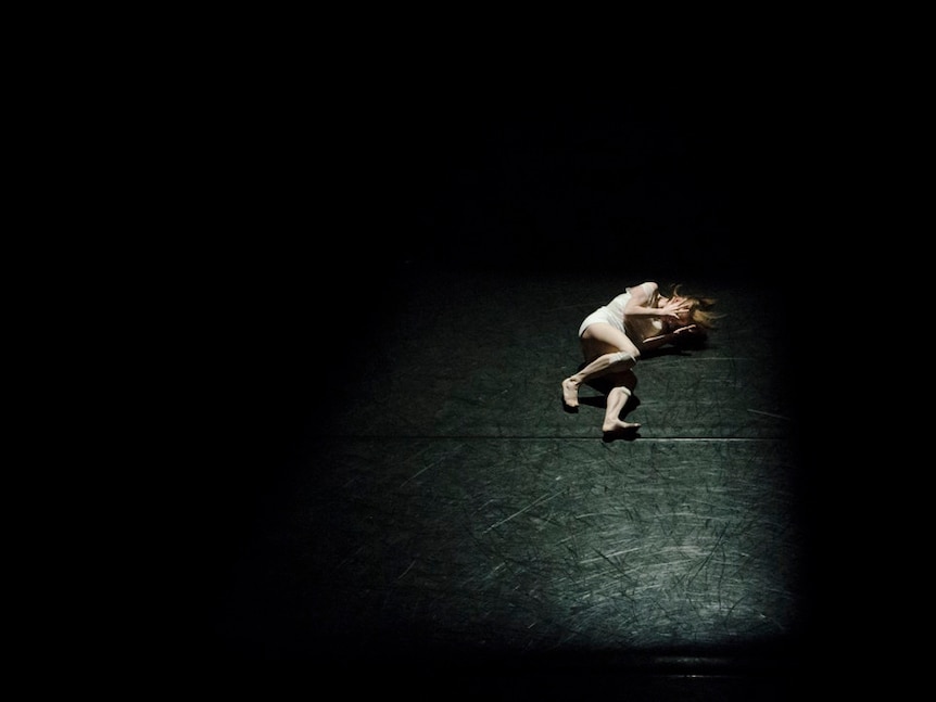 A woman in undergarments with tensed hands covering her face lay on a dark and empty black stage.