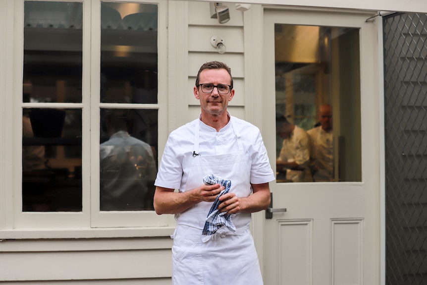 Chef wearing white uniform stands in front of outside wooden door and windows into kitchen