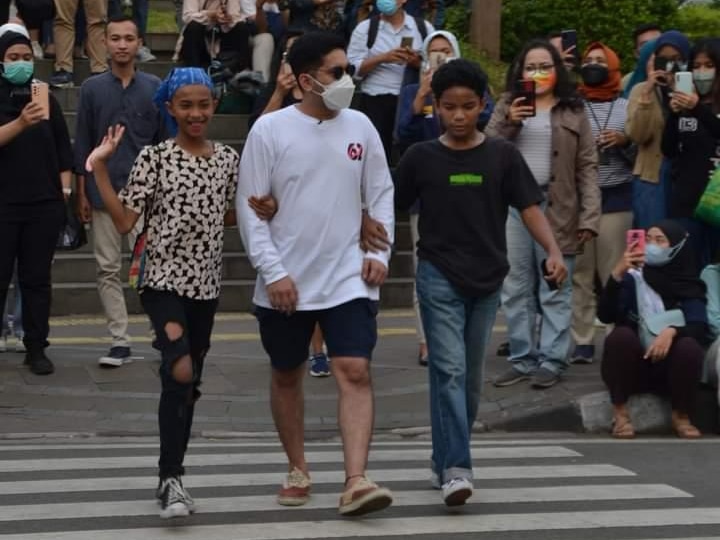Two teenagers wearing t-shirt and jeans and a man wearing long sleves shirt and short pants, walking in zebra crossing.