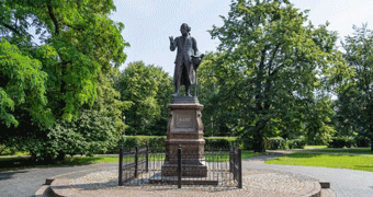 A statue of a man stands on top of a plinth in front of a green, leafy park.