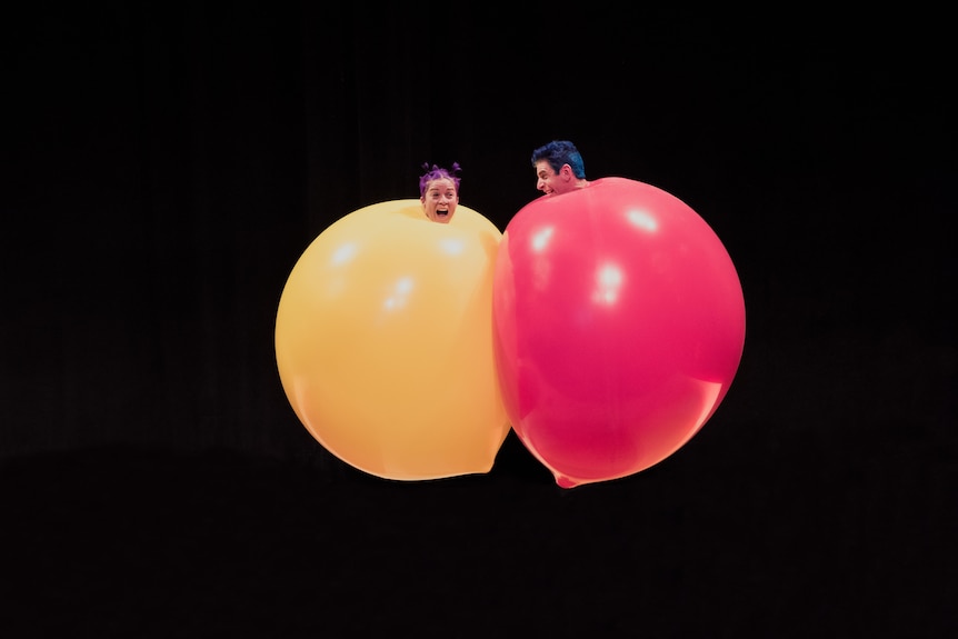 A woman inside a yellow balloon next to a man inside a red balloon, they are both in mid-air on a black background
