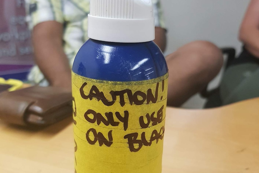 Blue bottle with white lid and wrapped in yellow tape with the words 'CAUTION! ONLY USE ON BLACKS' written on it.
