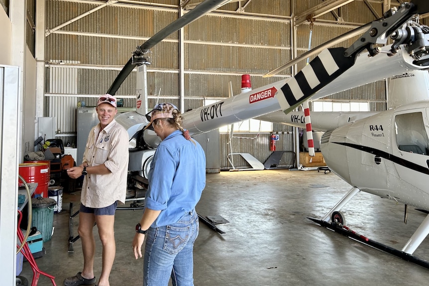 Smiling man in peach shirt, shorts, woman in blue shirt, jeans, blonde pigtail, cap, head bowed, next to a helicopter in hangar.