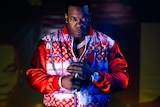 Busta Rhymes wears a half-zipped red jacket, a big chain, ring and watch. His hands are together. He stares at the camera.