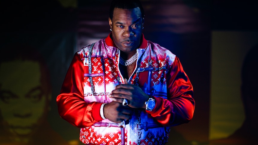 Busta Rhymes wears a half-zipped red jacket, a big chain, ring and watch. His hands are together. He stares at the camera.
