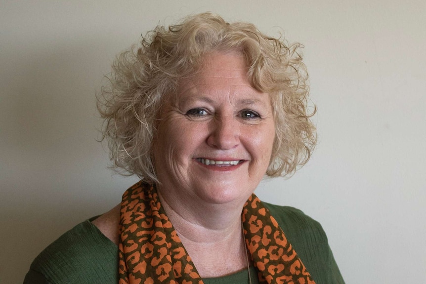 A smiling woman stands in front of a white wall smiling and wearing a green top with a black and yellow scarf