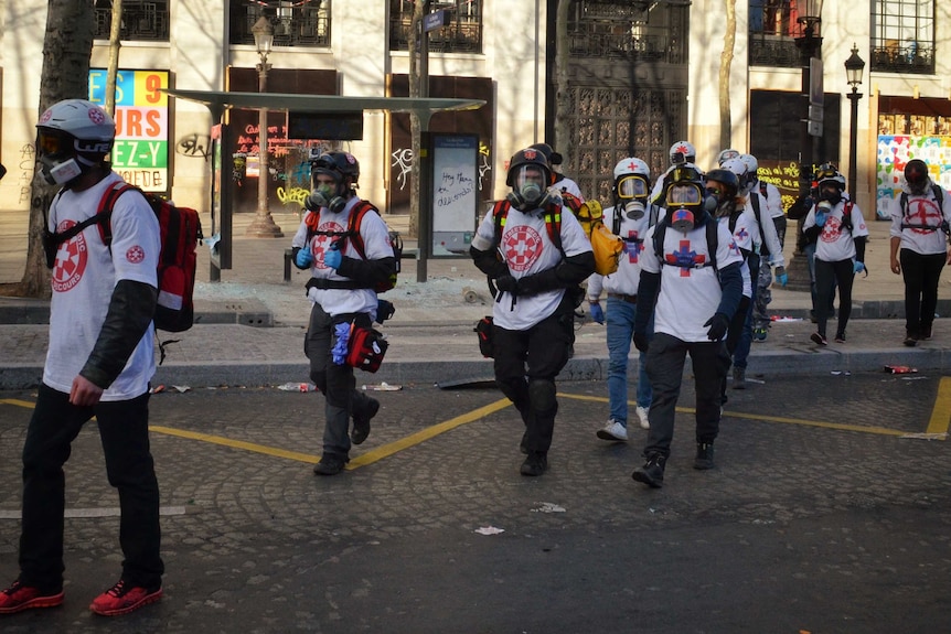 A group of medics wearing gas masks walk through a street past a smashed bus stop.