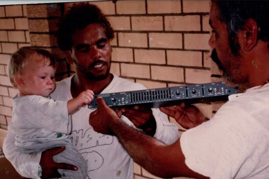Blurry family photo of two man playing with musical amplifier and small child on father's hip.
