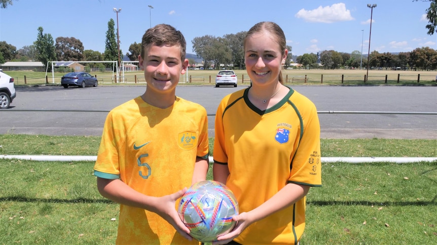A teenage boy and girl wearing gold Socceroos jerseys and holing a soccer ball in their hands