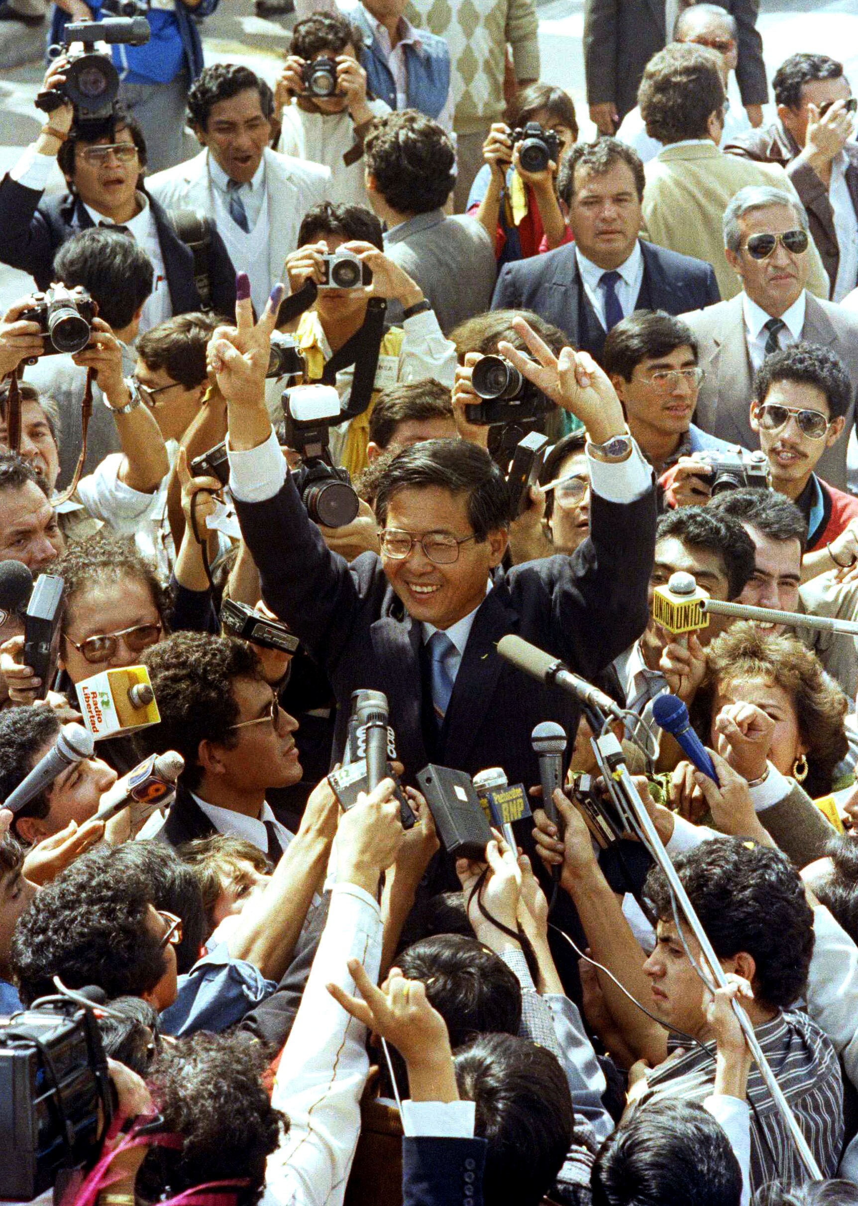 Alberto Fujimori surrounded by people holding microphones, with his hands in the air.