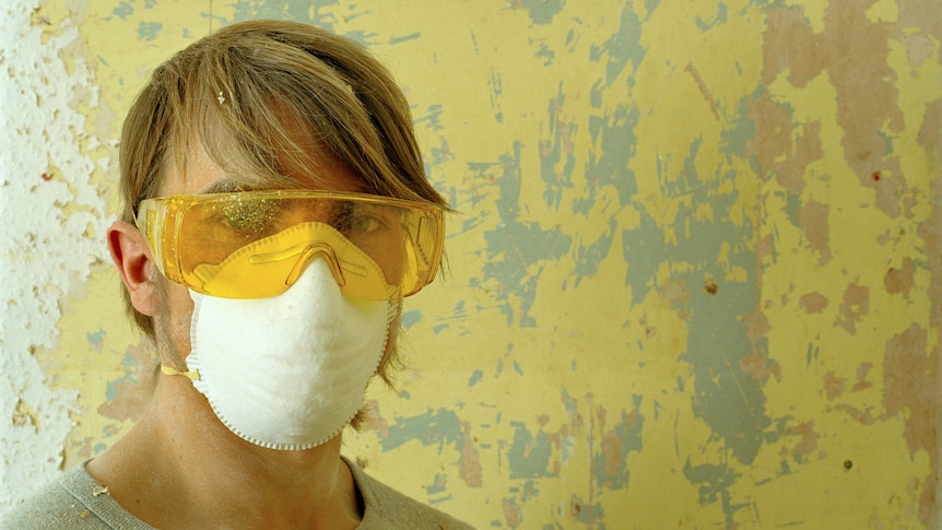 Against a yellow peeling paint background, a person with short fair hair wears industrial sized glasses and a white face mask. 
