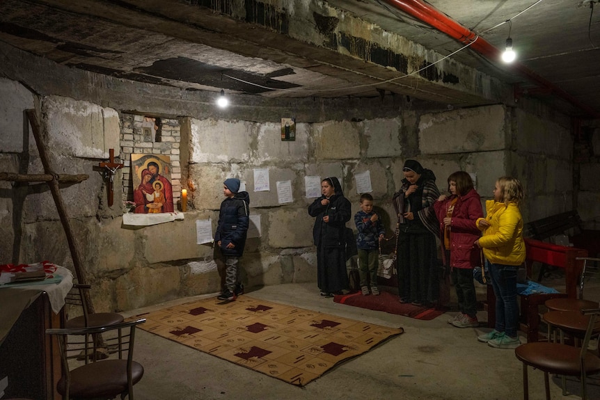 Children and a nun stand in a makeshift chapel in a basement, looking at a large crucifix and religious painting.