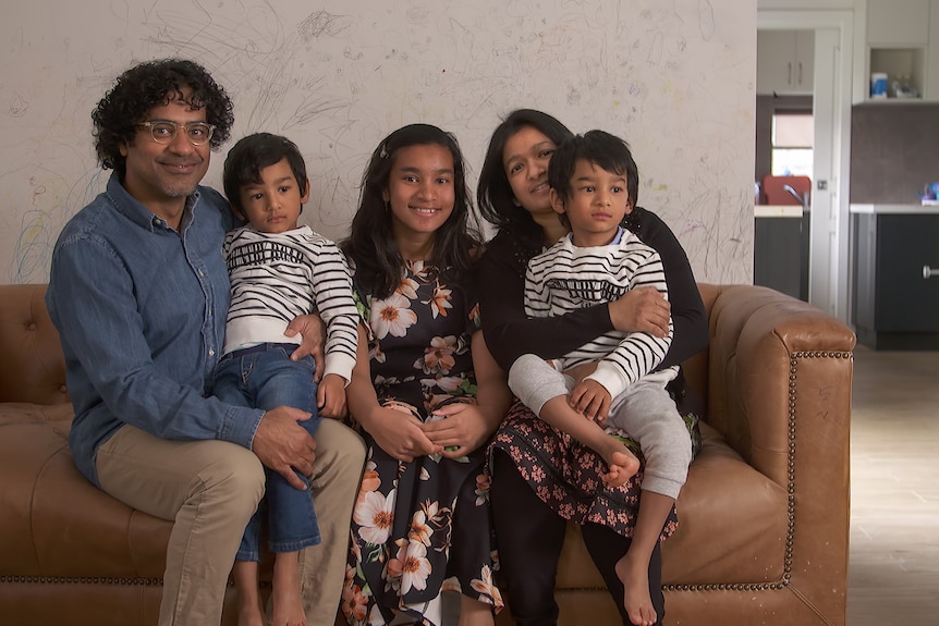 A mum and dad with their three kids sit on a brown couch smiling at the camera