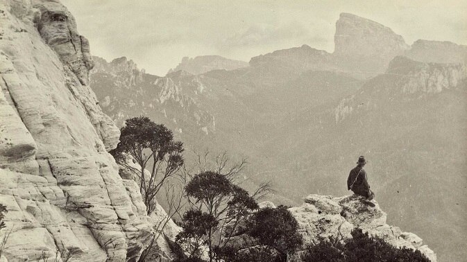 Black and white photo of a man sitting on a rock up high with mountains in the background