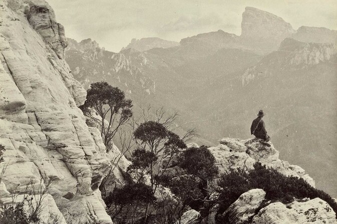 Black and white photo of a man sitting on a rock up high with mountains in the background