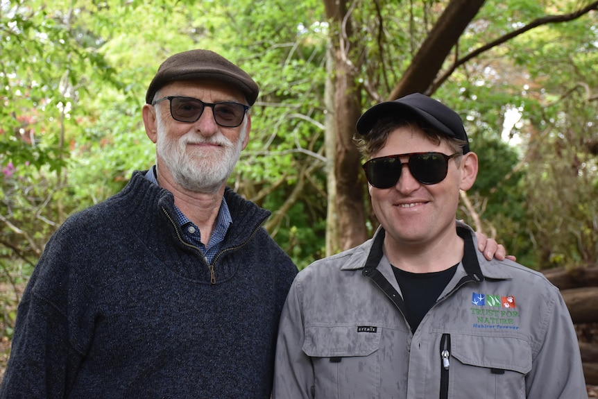 Two smiling men standing together with trees in the background.