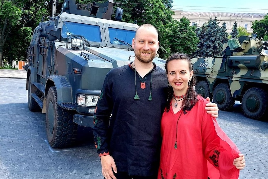 A man and woman in traditional Ukrainian garb pose for a photo near military vehicles. 