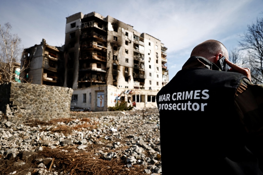 A man with a jacket labelled 'war crime prosecutor' talks on the phone with back to camera and destroyed buidings in background 