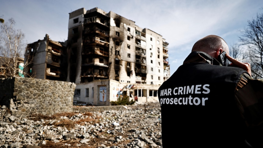 A man with a jacket labelled 'war crime prosecutor' talks on the phone with back to camera and destroyed buidings in background 