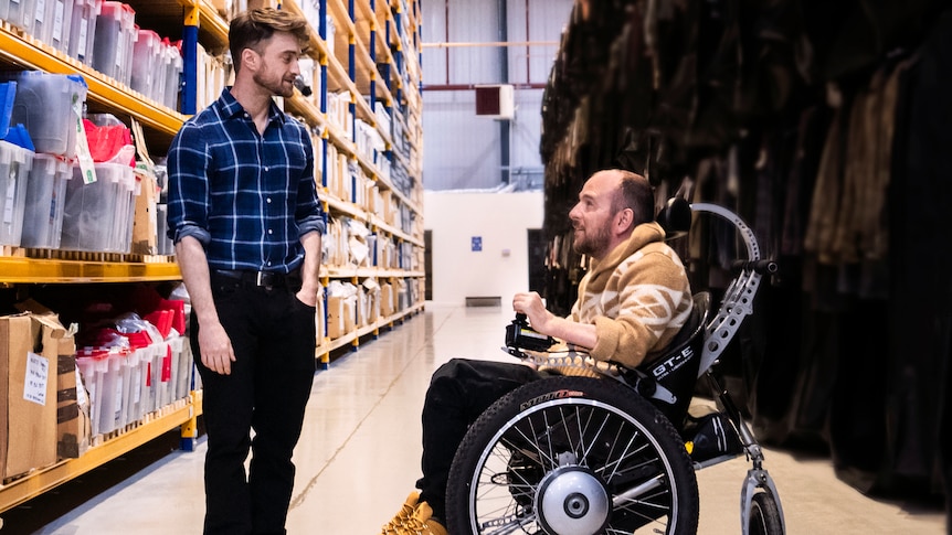 David, in a wheelchair, speaks to Daniel in a warehouse as Harry Potter costumes and props surround them.