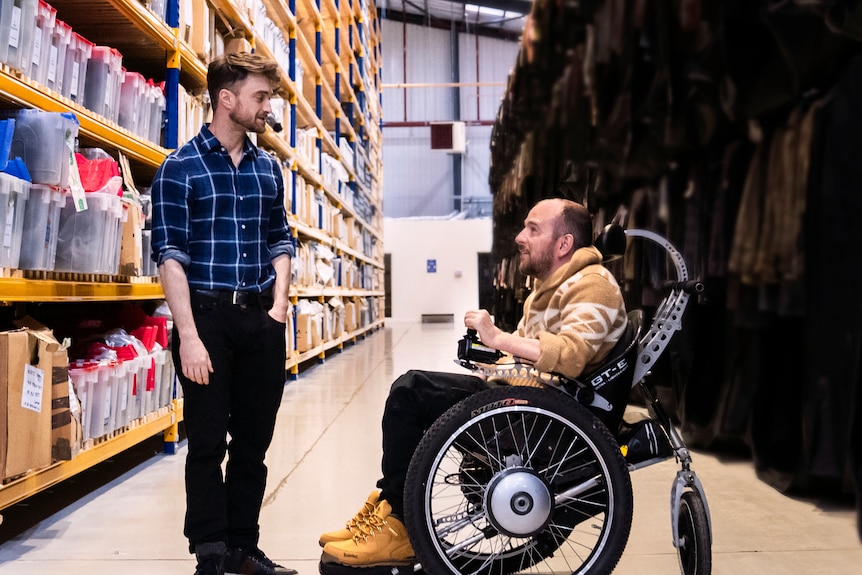 David, in a wheelchair, speaks to Daniel in a warehouse as Harry Potter costumes and props surround them.