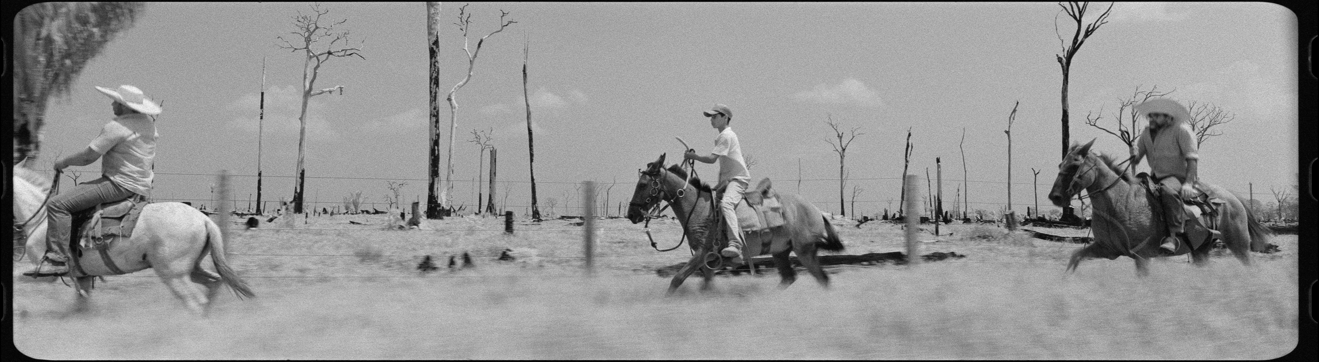 A black and white still from a video artwork featuring three men in cowboy hats riding horses through a barren landscape.