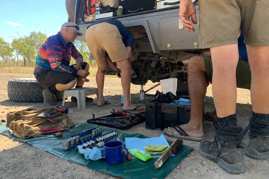 Tools on the ground and a vehicle with its wheel off, and four men looking on.