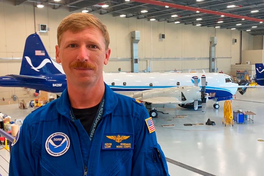 A man with a neat moustache wears a blue shirt and stands in front of a plane inside a hangar.