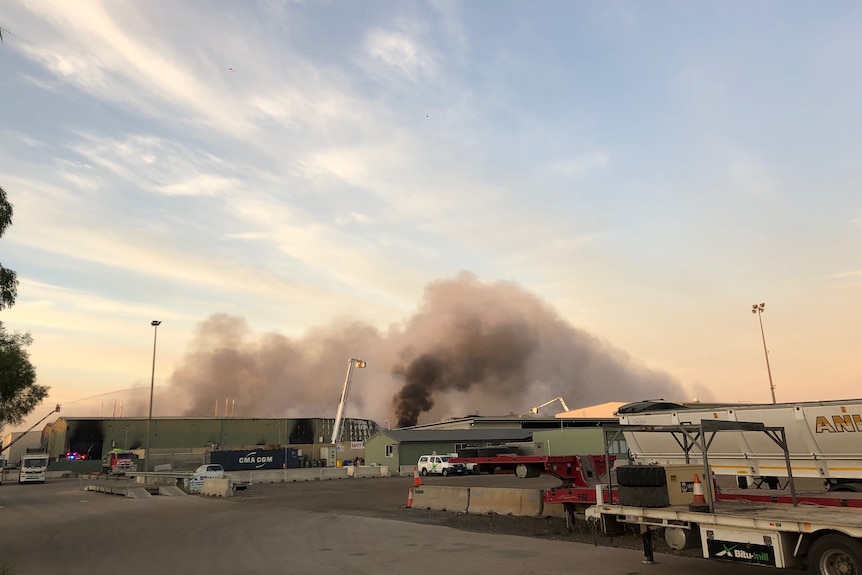 Fire fighting cranes aim water at a factory fire which is billowing smoke over the horizon