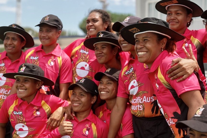 A team of female cricketer in red and black, wearing broad-brimmed black hats, huddling together and smiling.