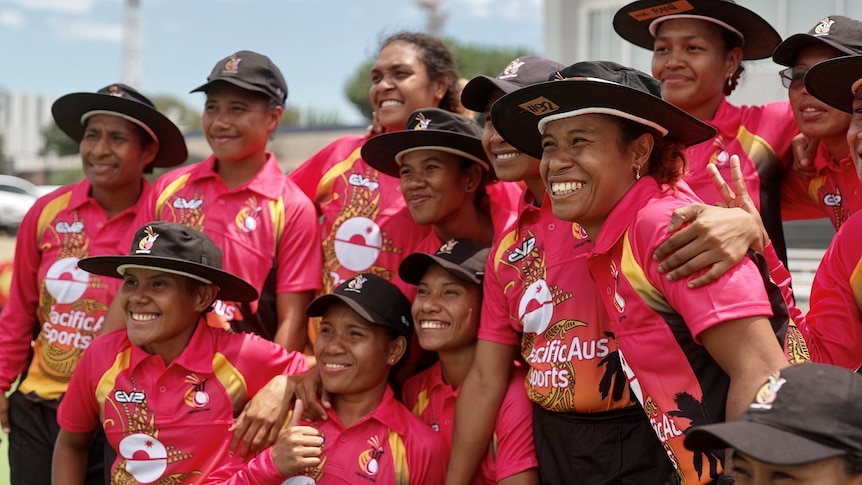 A team of female cricketer in red and black, wearing broad-rimmed black hats, huddle together and smile for a photo.