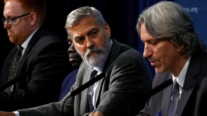 A bearded man wearing a suit sits between two men at a table, and turns to talks to one of them.