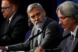 A bearded man wearing a suit sits between two men at a table, and turns to talks to one of them.