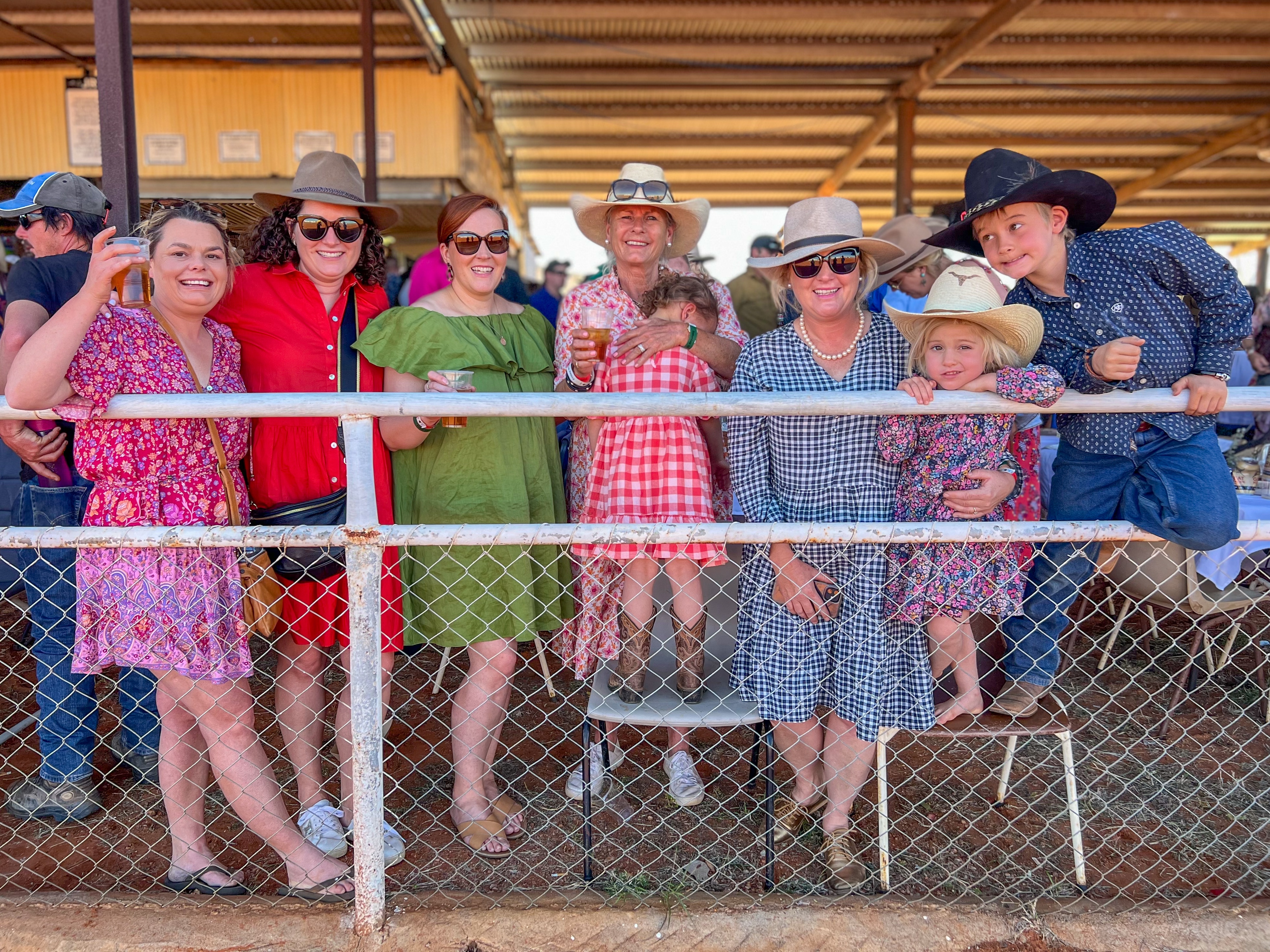 A group of women and children line the horse racing track, smiling at the camera