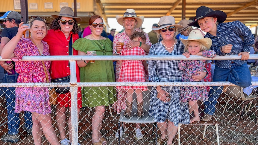 A group of women and children line the horse racing track, smiling at the camera