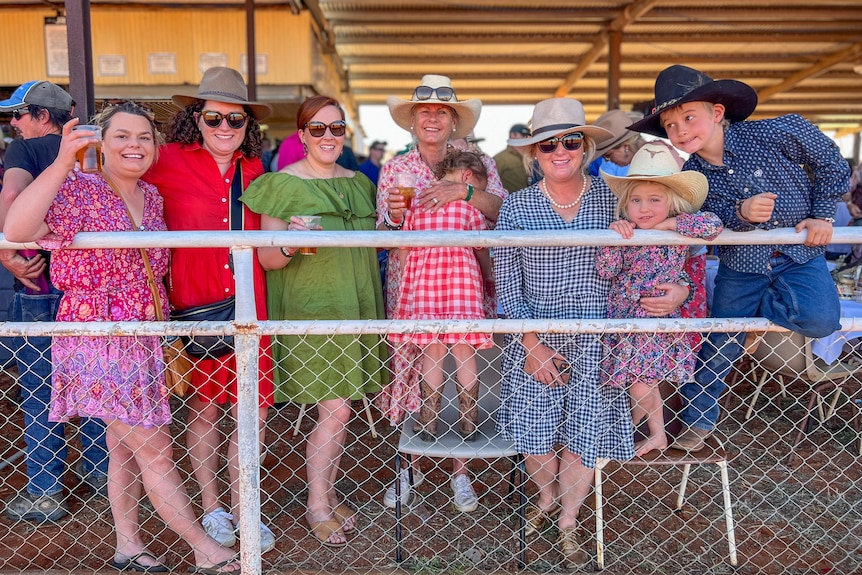 A group of women and children line the horse racing track, smiling at the camera