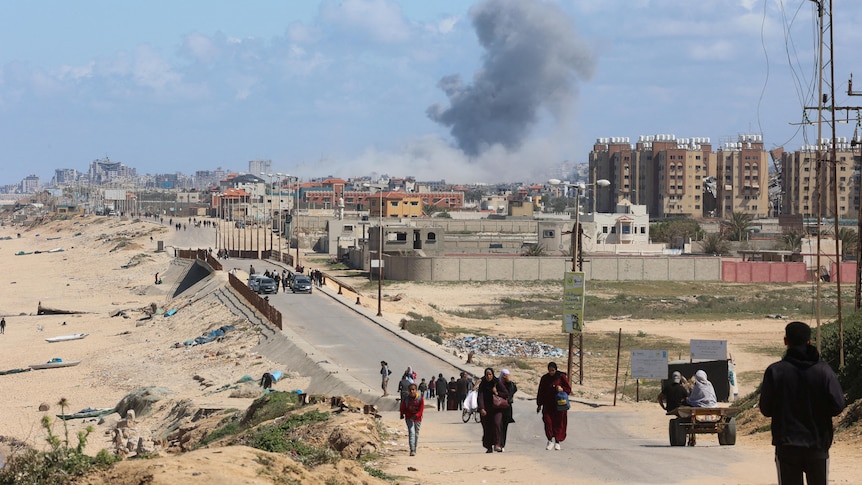 Palestinian civilians walk along a long road as a smoke cloud rises from buildings in the background.